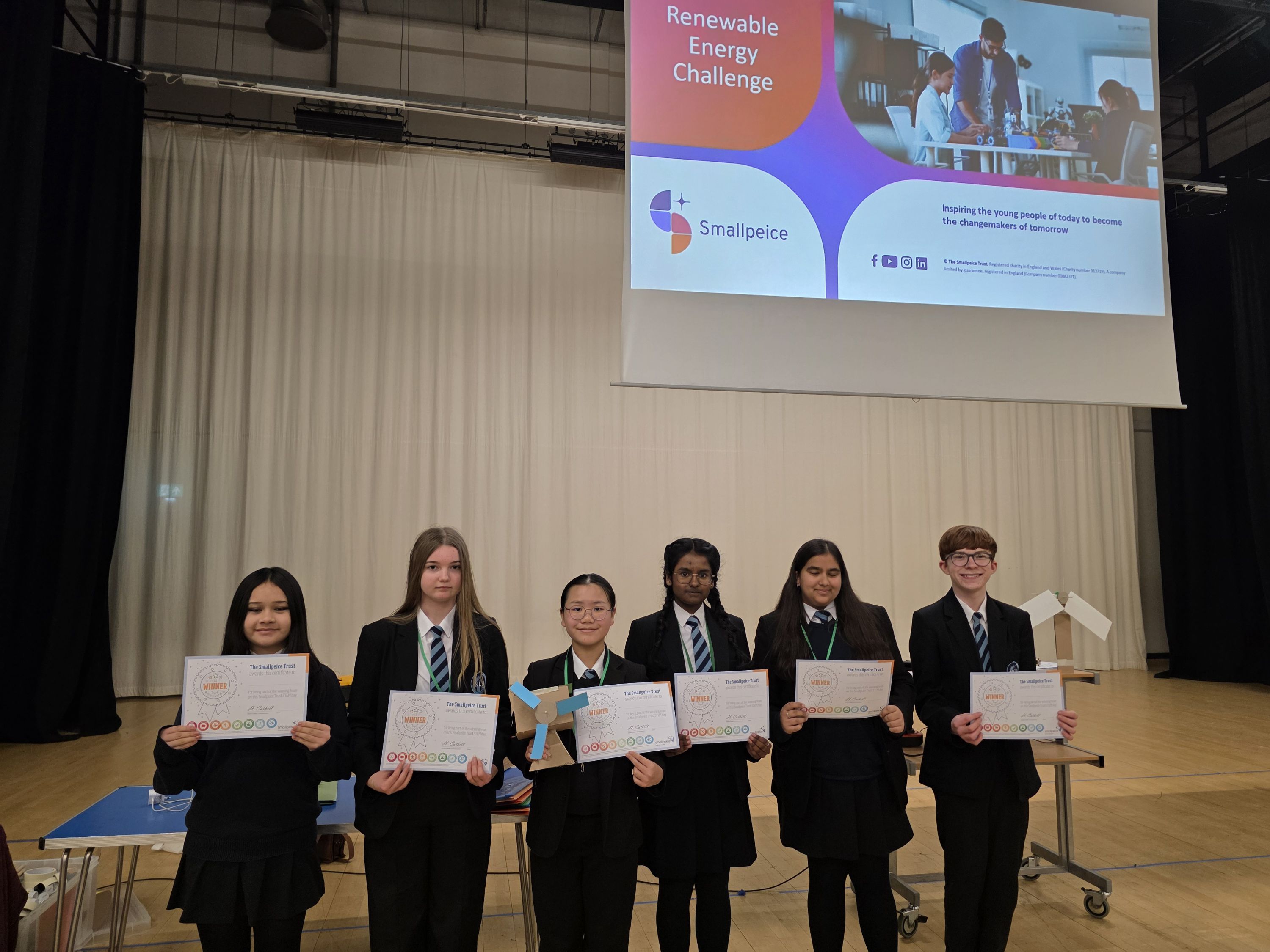 Photograph of six students standing  holding certificates and a small wind turbine model, participating in a Renewable Energy Challenge. A presentation slide above them displays the challenge title, Smallpiece logo, and supporting text about encouraging young people in engineering and technology.