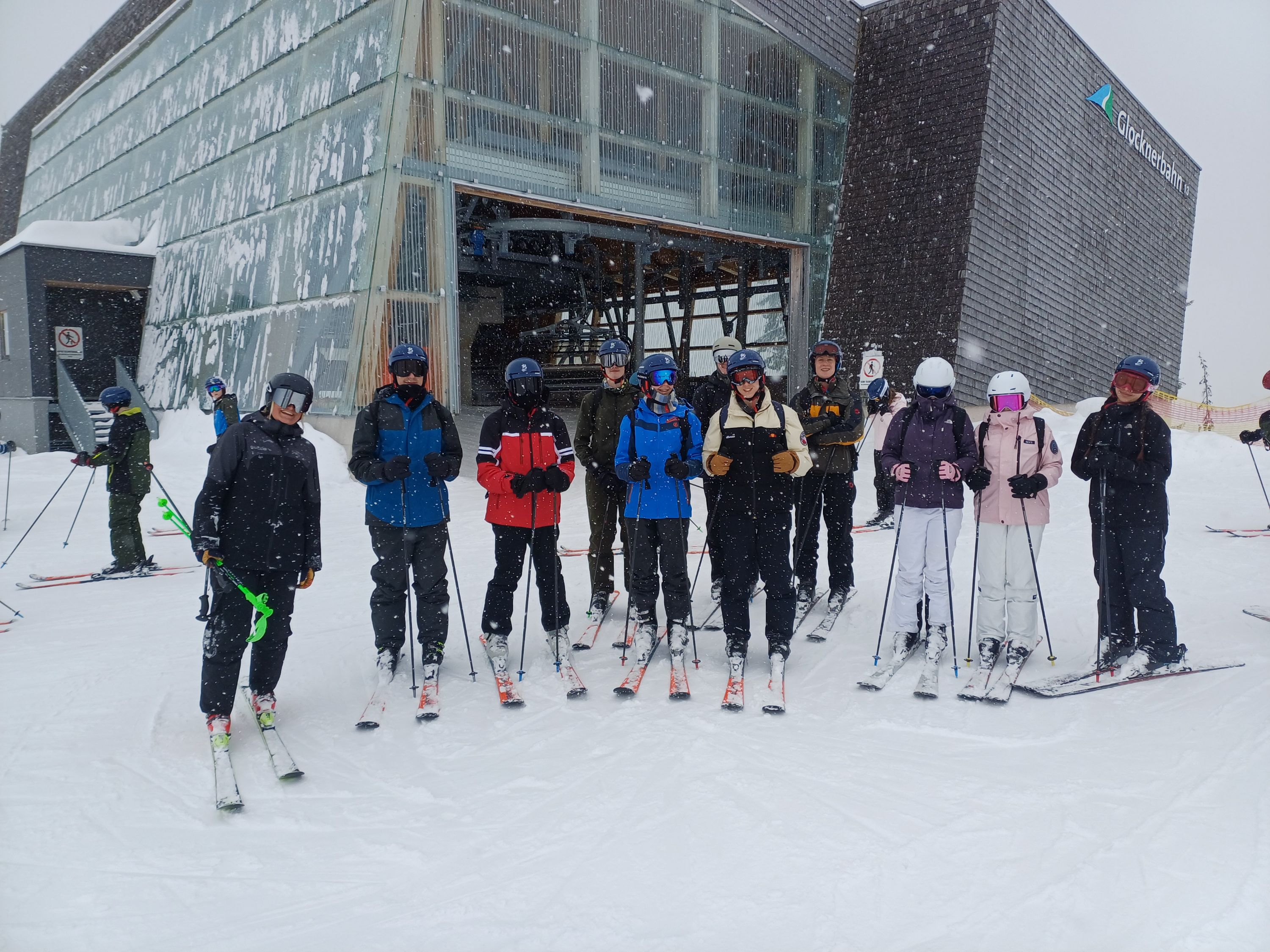 Photograph of a group of ten skiers standing on snow in front of a modern ski lift station during snowfall. Skiers wear various coloured jackets and helmets, holding ski poles, with some people skiing in background.