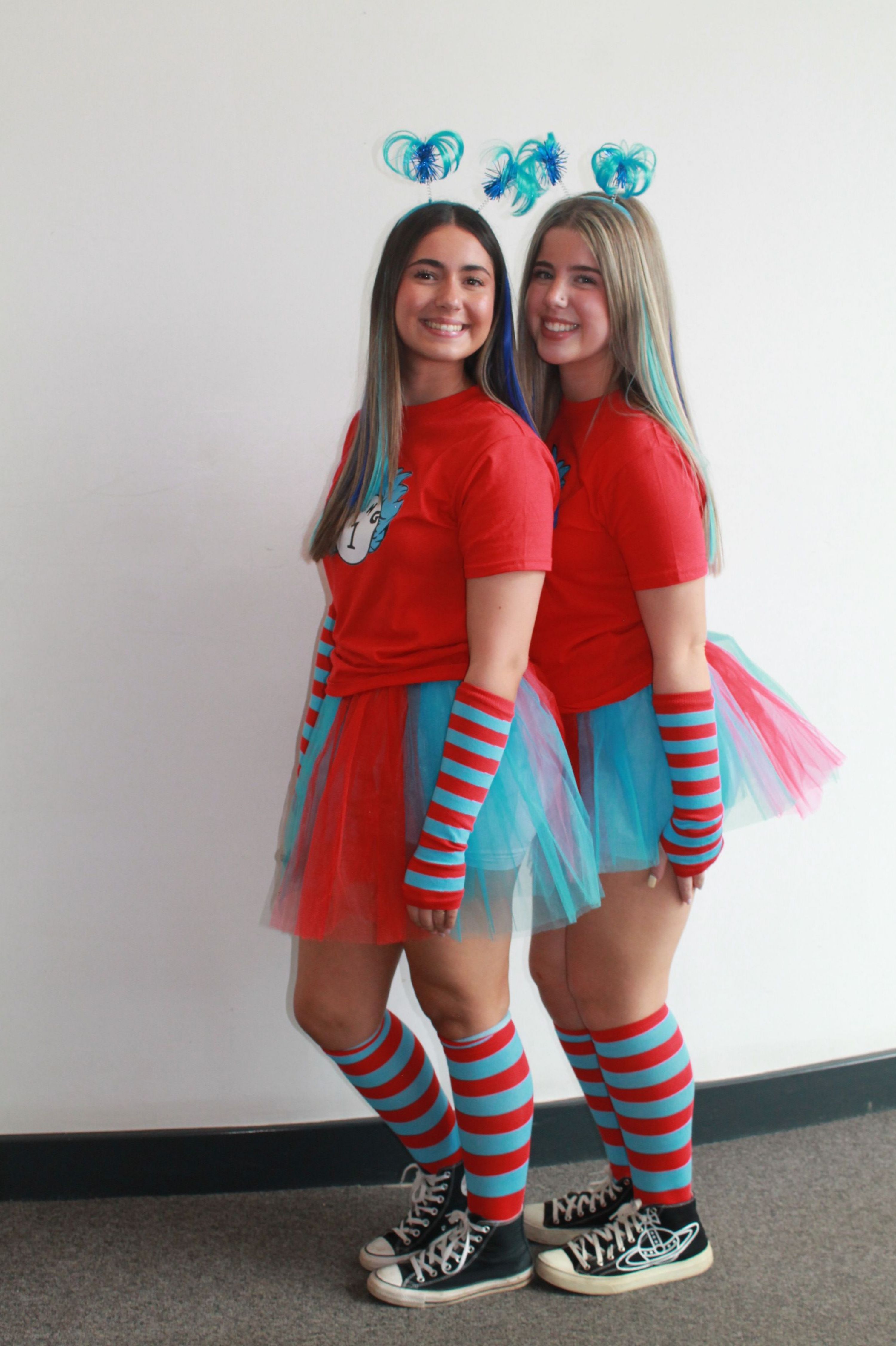 Photograph of two women dressed in matching colourful costumes featuring red tops, blue and red striped arm warmers and knee-high socks, blue tutus, and black Converse shoes. Both have long hair with blue streaks and are standing side by side