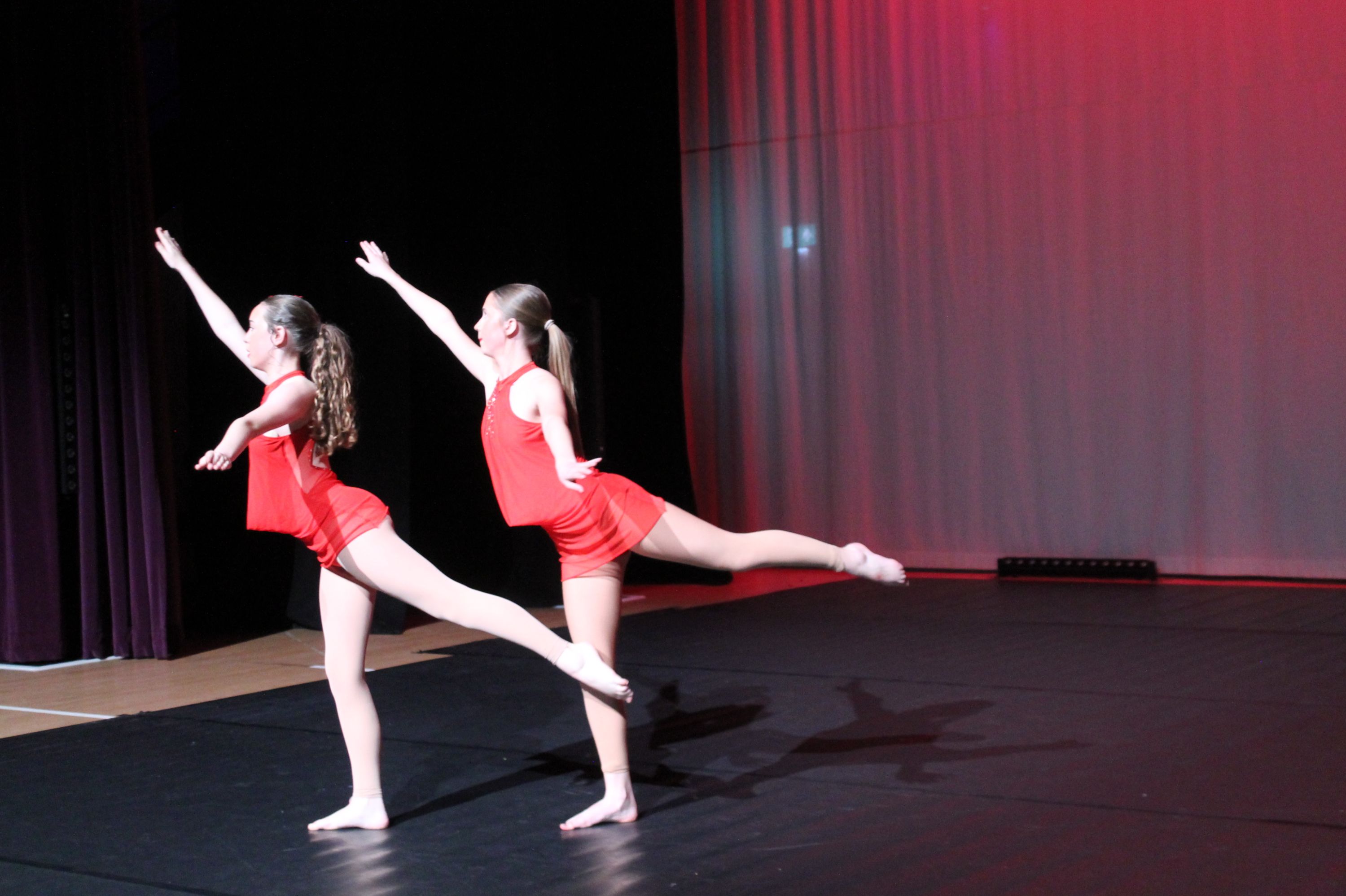 Photograph of two young female dancers performing a ballet pose on stage, both wearing red leotards and beige tights. The background features dark curtains on one side and a red-lit backdrop, highlighting their extended arms and legs in a balanced arabesque position.