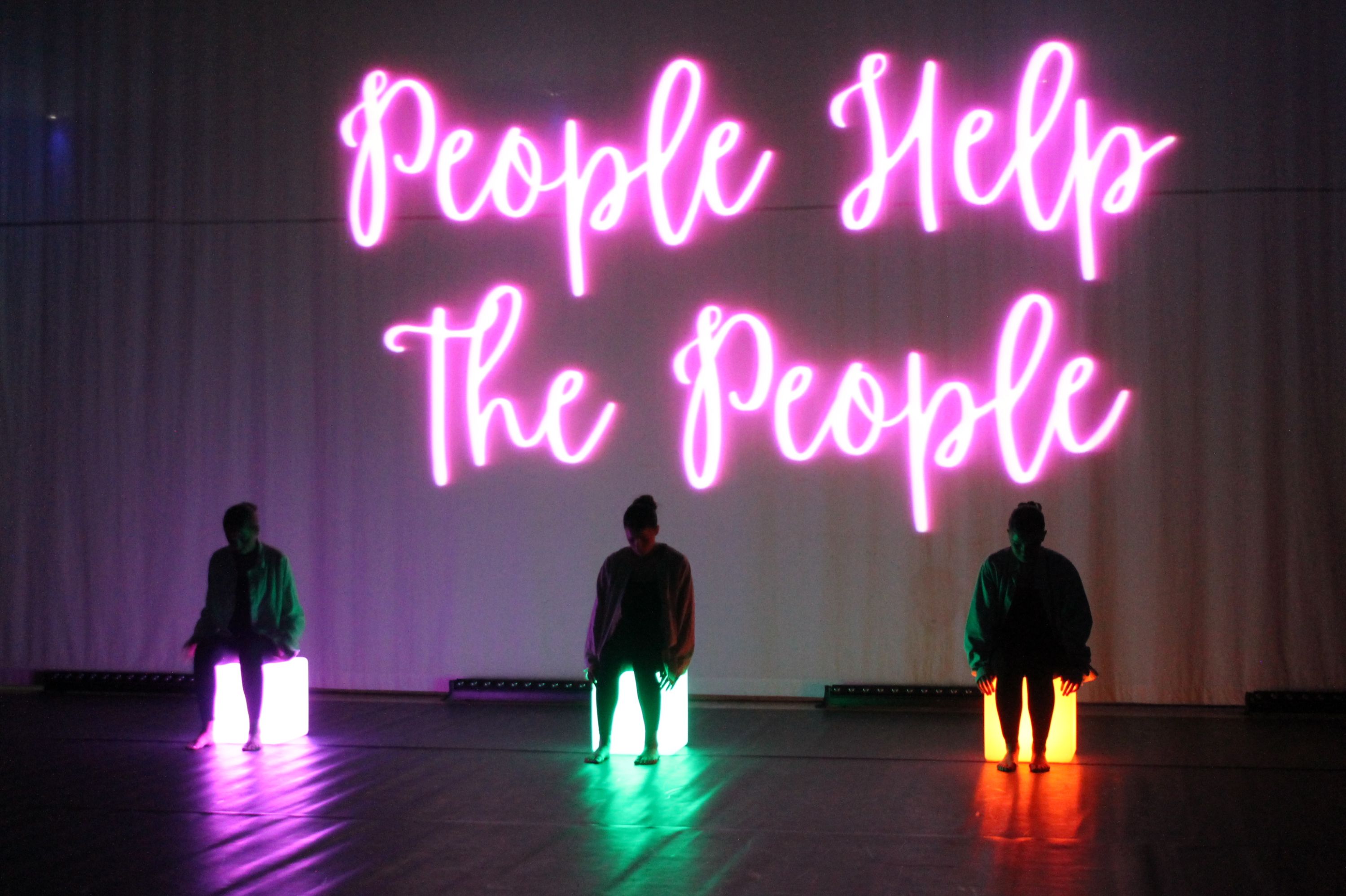 Photograph of three people sitting on illuminated cubes in a dark room with a large neon sign reading "People Help The People" in bright pink cursive letters behind them. The cubes emit different colours—white, green, and orange—creating a visually striking contrast against the dark floor and backdrop.