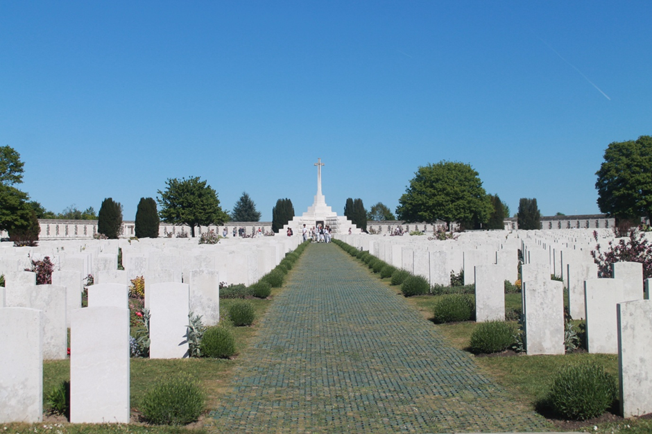 Photograph of Tyne Cot war cemetery featuring rows of white headstones aligned along a central paved pathway leading to a large white cross monument. The scene is set under a clear blue sky with green trees bordering the cemetery, highlighting a solemn and orderly memorial space.