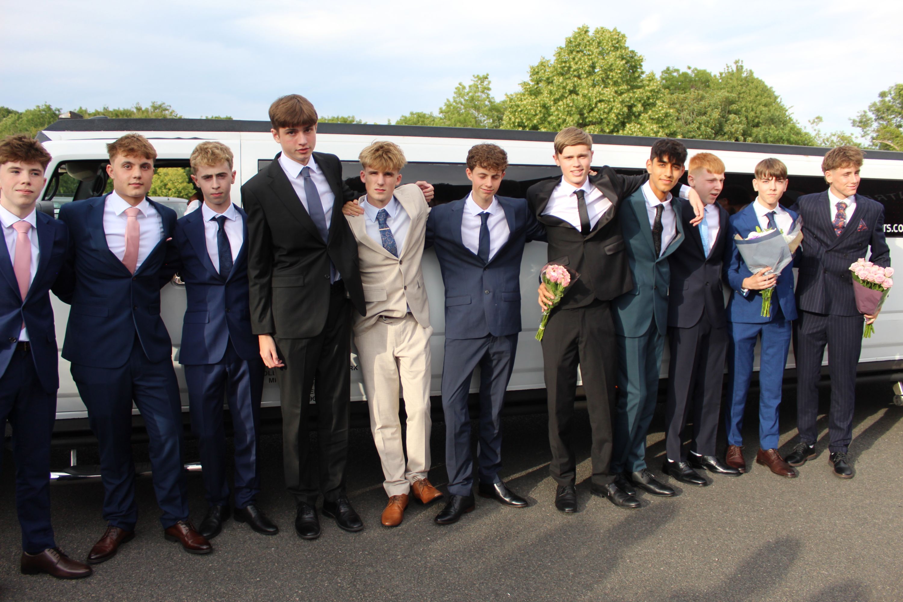 hotograph of a group of young men dressed in formal suits standing side by side in front of a white limousine, likely preparing for a special event such as prom or a formal celebration. Several hold bouquets of flowers, and the group displays a mix of dark, light, and patterned suits with ties, set against a backdrop of trees and a cloudy sky.