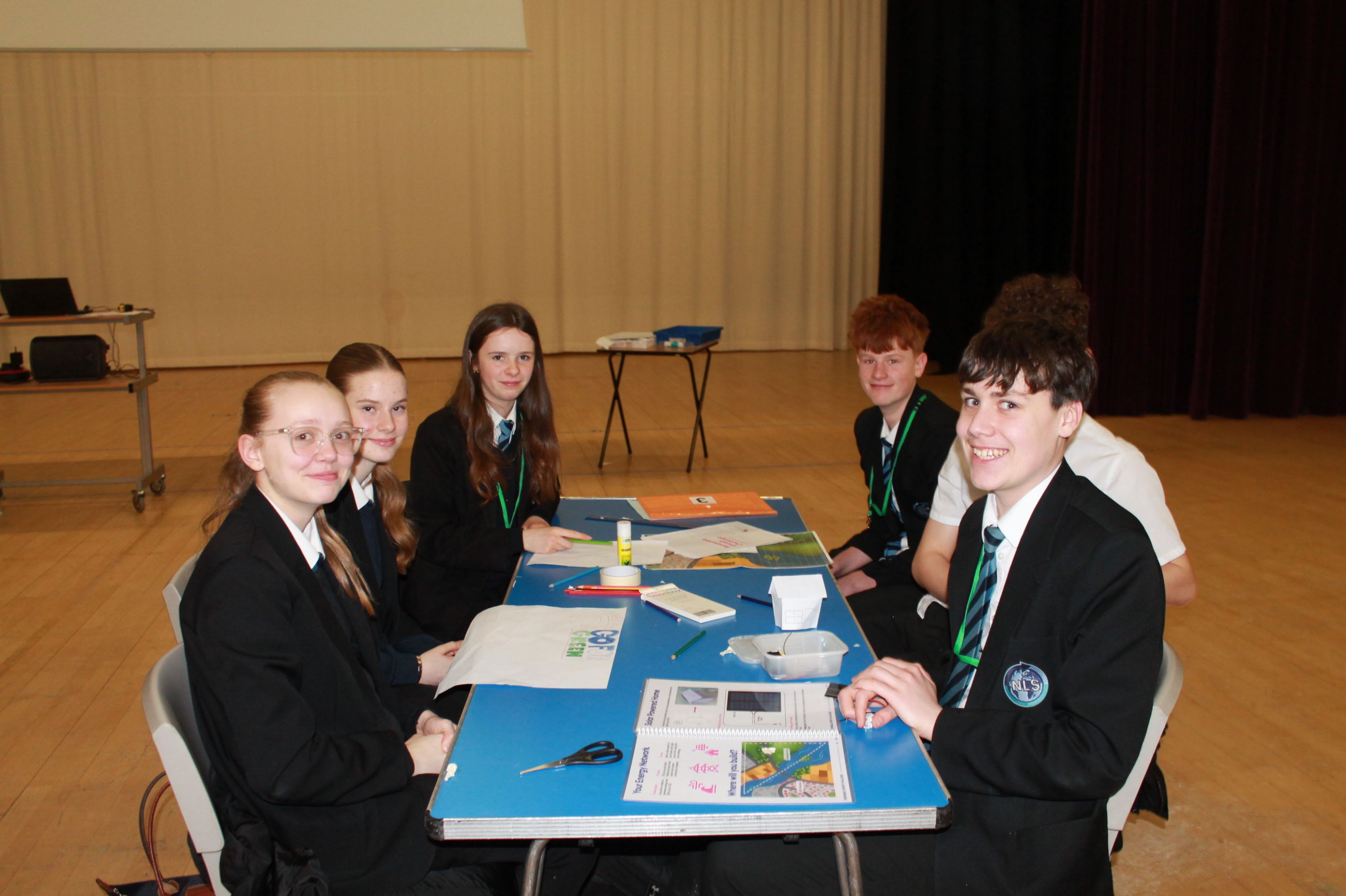 Photograph showing a group of six students in school uniforms seated around a blue table in a large room, engaged in a collaborative activity involving papers, glue sticks, and scissors. The setting appears to be an 