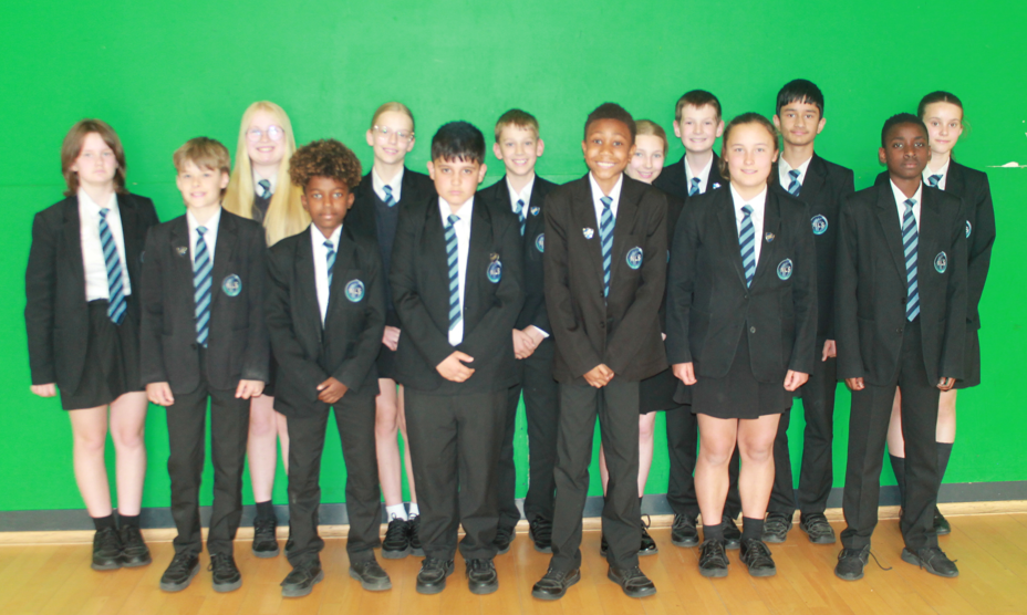 Photograph of a group of schoolchildren standing in two rows against a green wall, all wearing black blazers, white shirts, and striped blue and white ties as part of their uniform. The group includes both boys and girls, with some wearing skirts and others trousers, positioned on a wooden floor indoors.