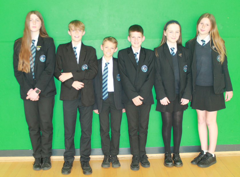 Photograph of six schoolchildren standing in a row against a green background, all wearing black blazers with school badges and striped ties. The group includes both boys and girls, with variations in skirt and trouser uniforms,