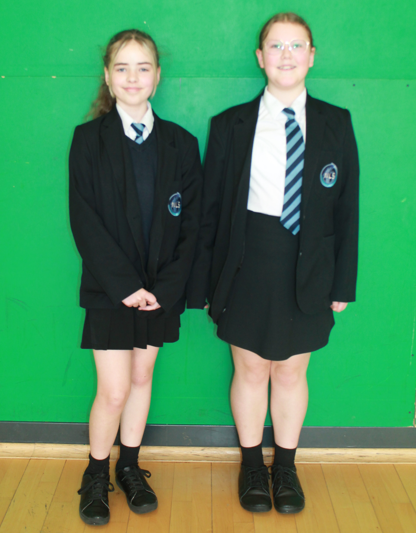 Photograph of two schoolgirls standing side by side against a green background, both wearing black blazers with school crests, white shirts, and striped ties. 