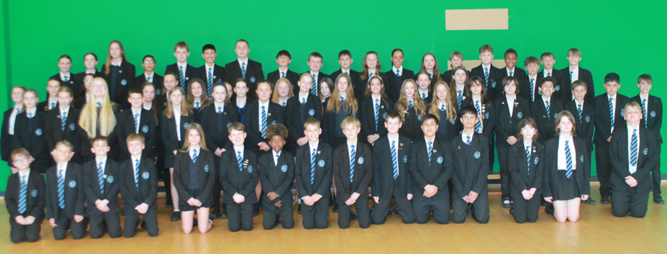 Photograph of a large group of school students in uniform posing indoors against a green wall, arranged in three rows with some standing and others kneeling. Students wear black blazers, white shirts, and striped blue ties, 