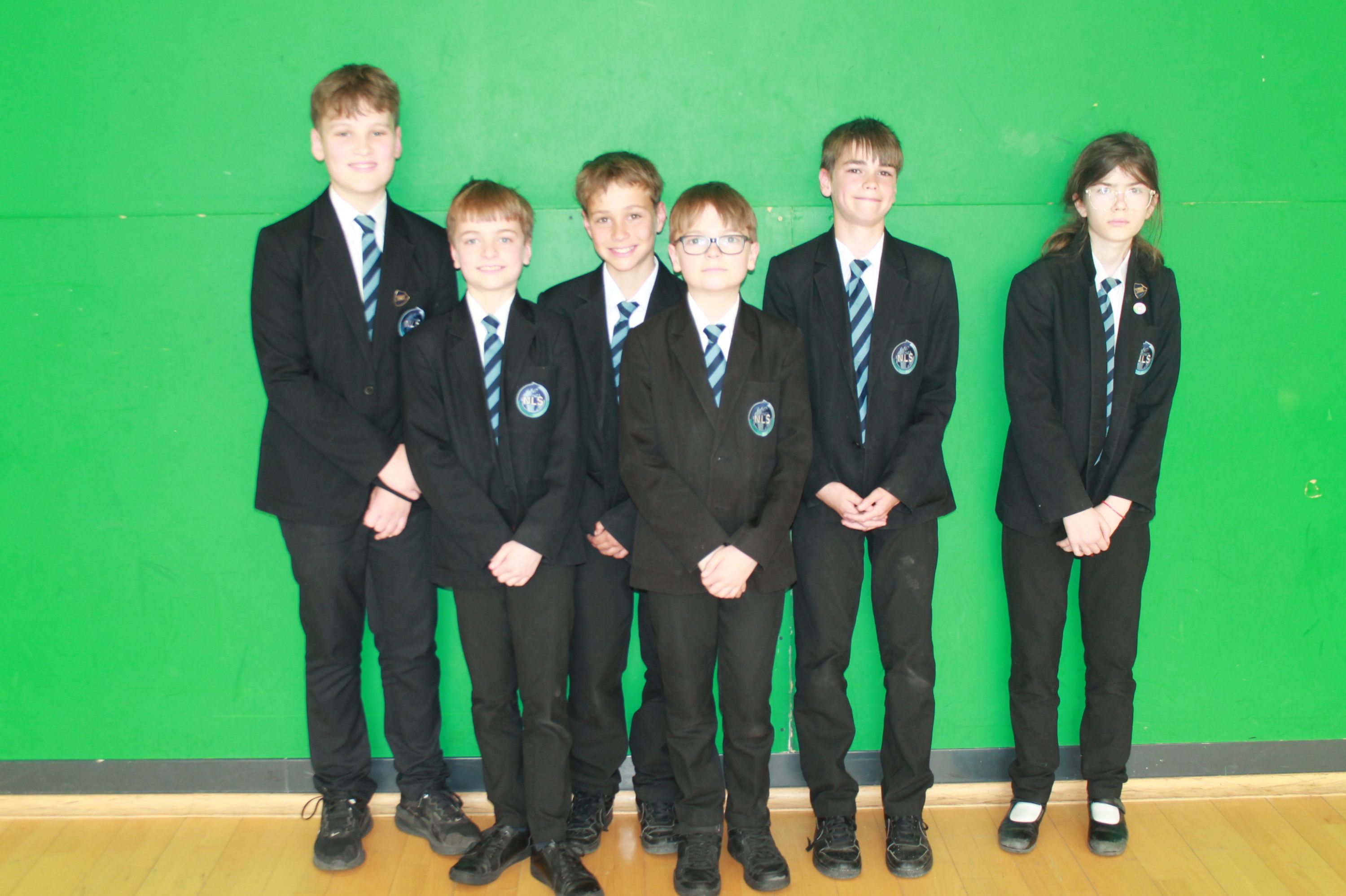 Photograph of six schoolchildren standing in a row against a green wall, all wearing black blazers, white shirts, and striped ties as part of a school uniform. The group includes both boys and girls, with all students facing forward and standing on a wooden floor.