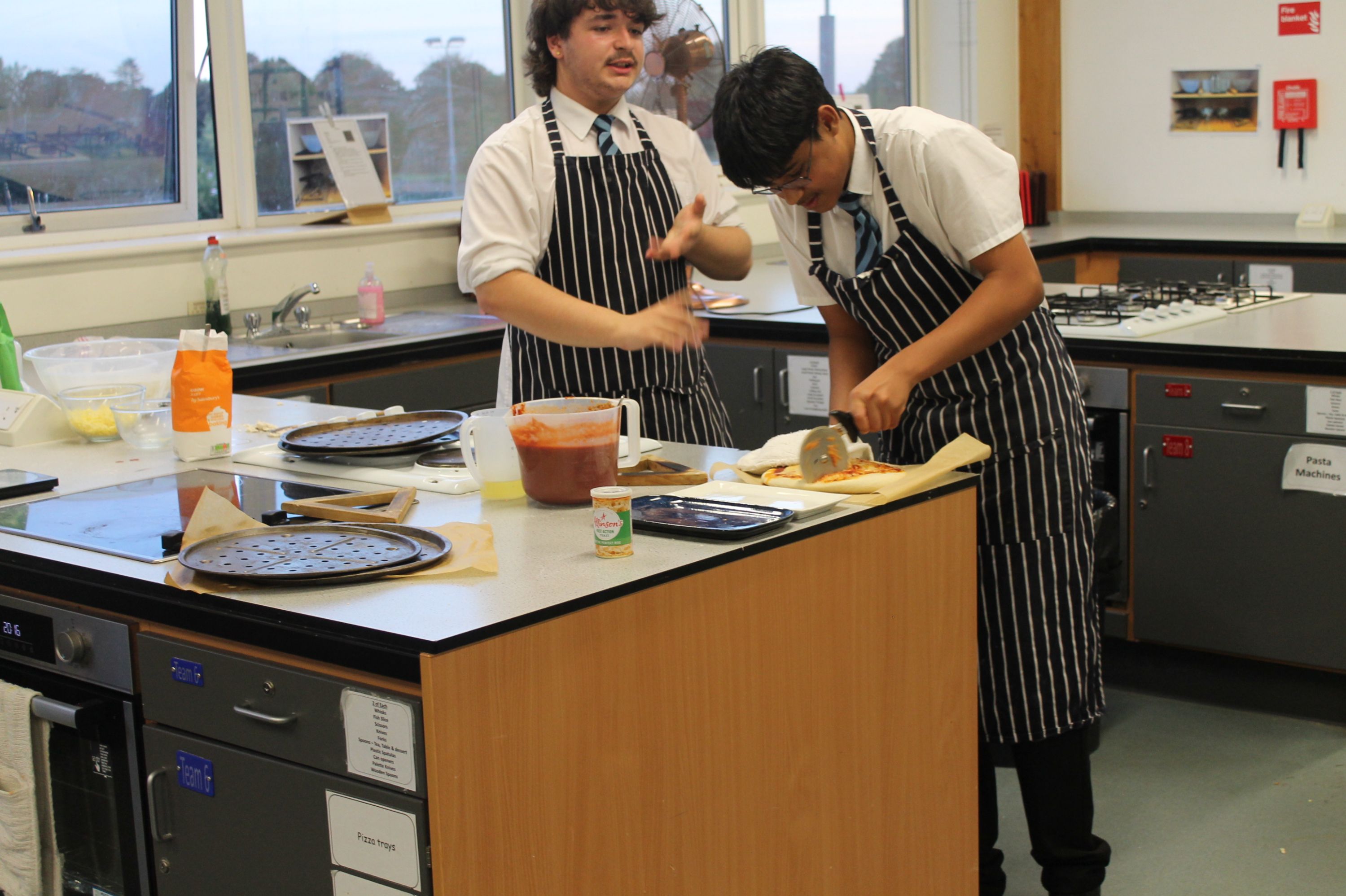 Photograph showing two students in striped aprons preparing pastry at a school kitchen workstation. Rolling trays, ingredients, utensils, ovens and labelled storage units indicate a practical cookery lesson.