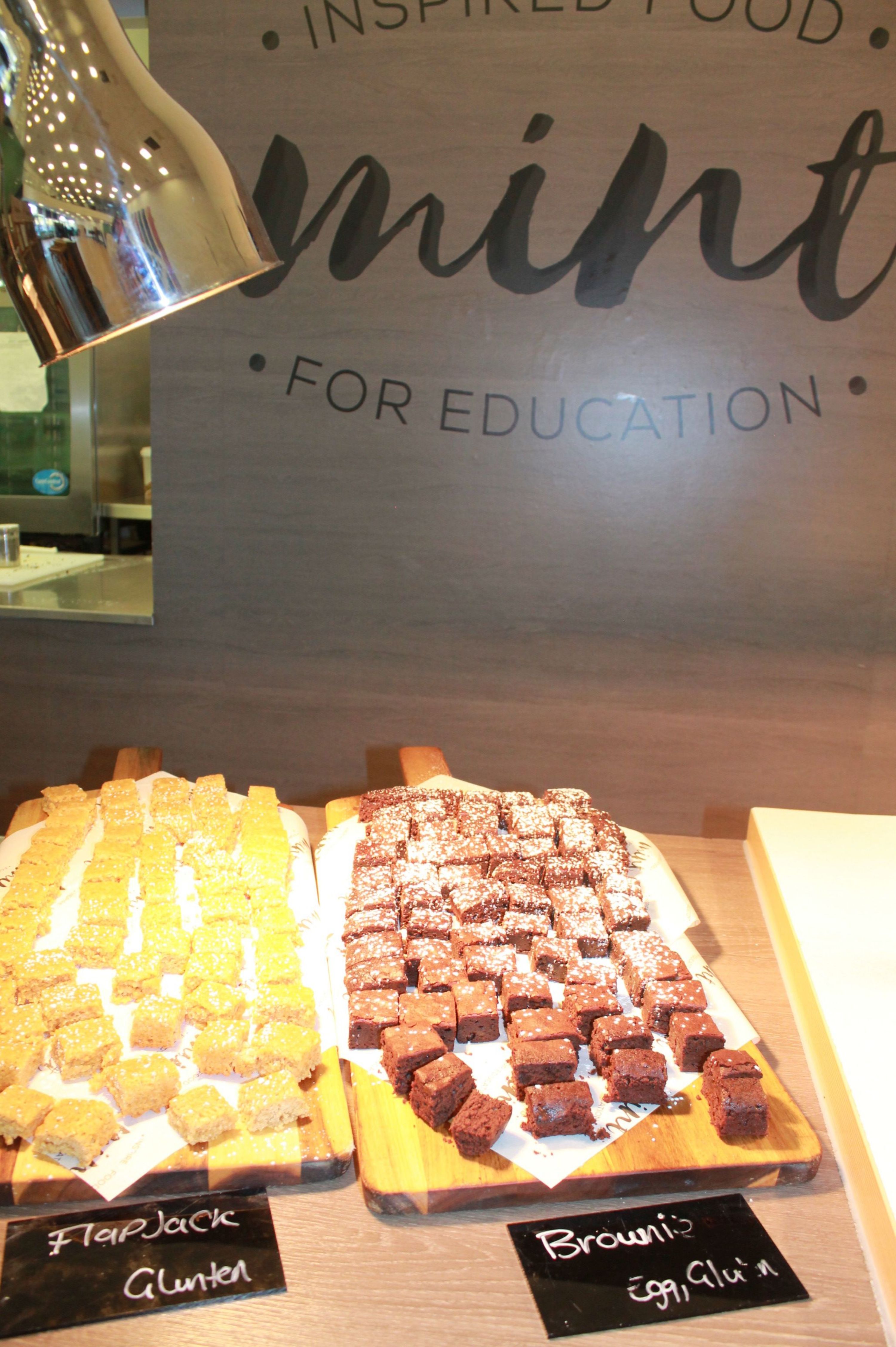 Photograph of  counter displaying trays of assorted square cakes and brownies. Warm lighting highlights chocolate and golden pieces, with black handwritten labels and background lettering reading “INSPIRED FOOD” and “FOR EDUCATION”.