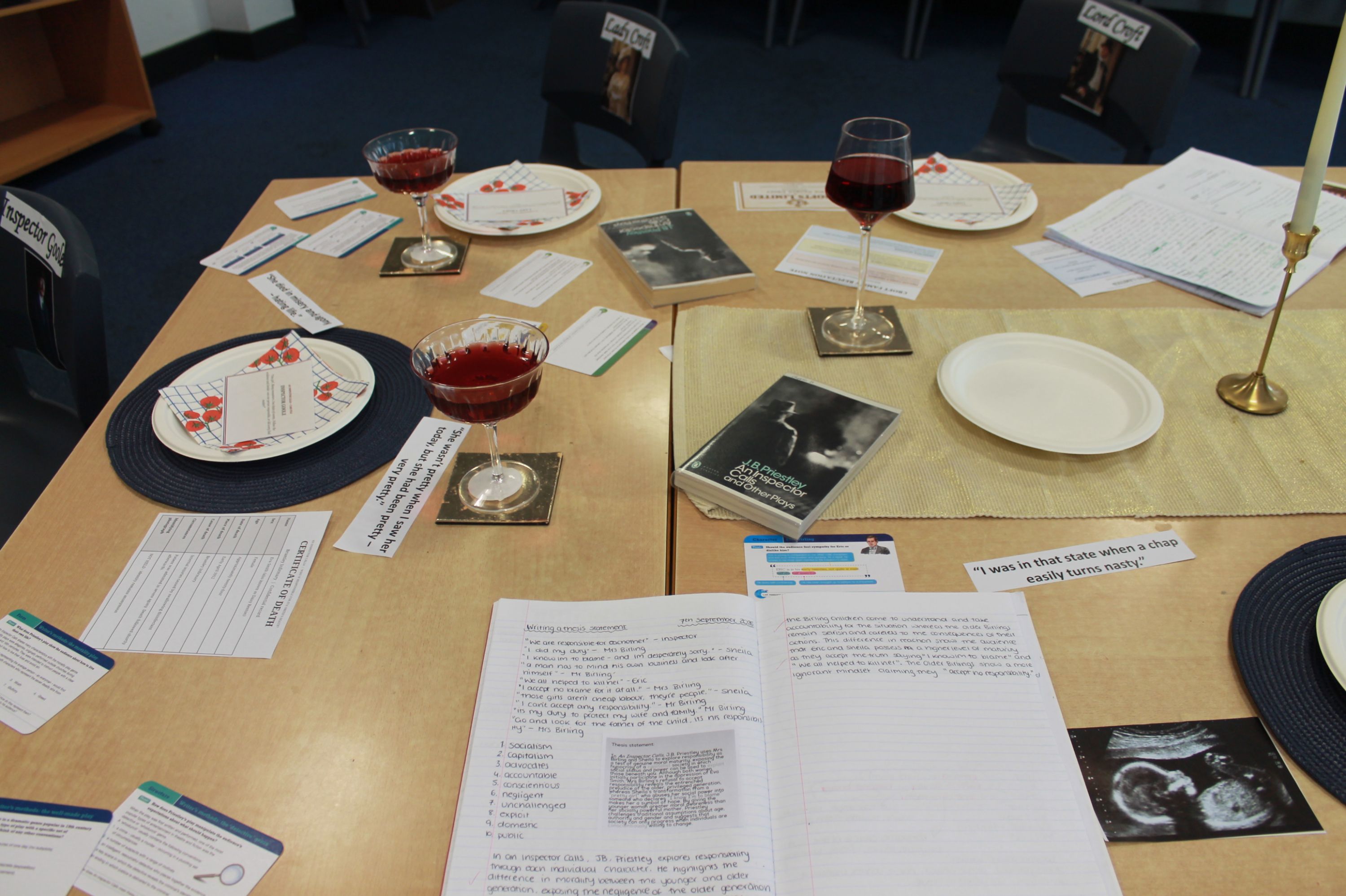 Photograph showing a staged dining-table display combining dinnerware, wine glasses, candles, printed forms, cards and open booklets for an educational or investigative activity. Medical ultrasound photographs, handwritten notes, questionnaires and labelled plates provide notable clues, while blue placemats and scattered documents structure each place setting.