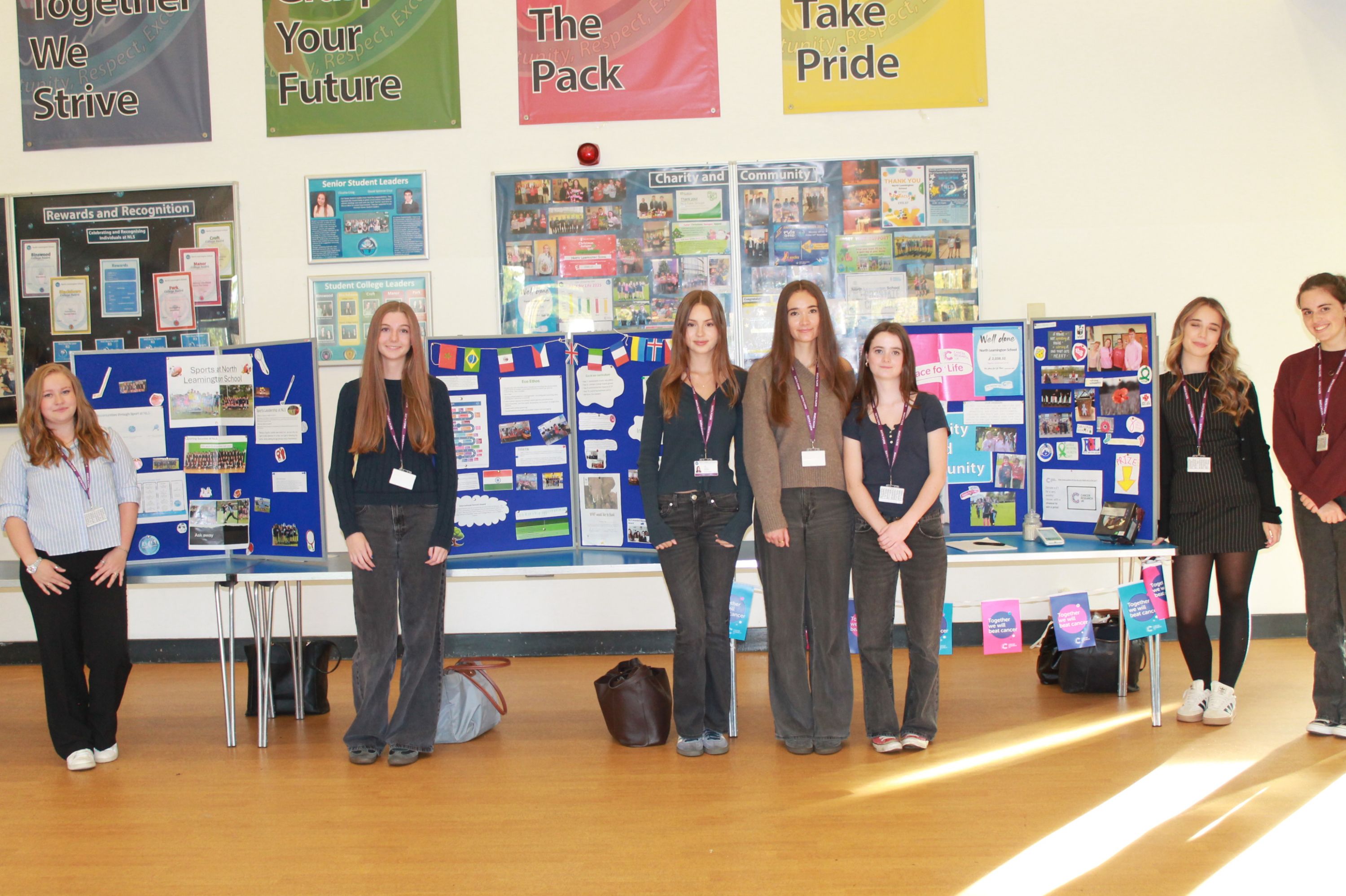 Photograph of students presenting colourful  display boards.  Posters, diagrams, photographs, certificates and wall banners surround presenters wearing identification lanyards, with sunlight across wooden flooring.
