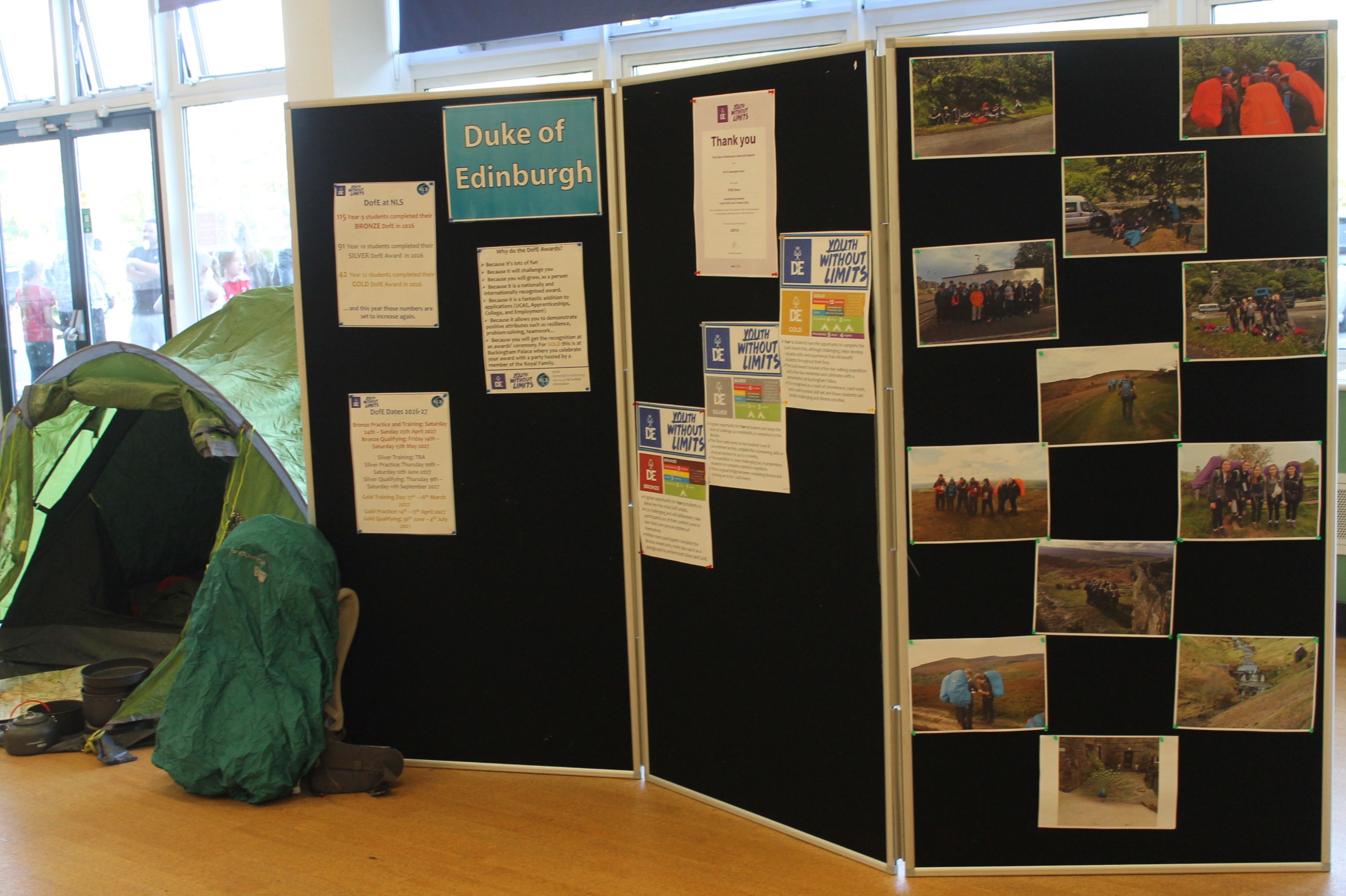Photograph of a Duke of Edinburgh award display board showing certificates, programme information, expedition photographs and participant groups. Black panels feature blue-and-yellow award labels, while a green tent and outdoor equipment highlight expedition activities and achievement milestones.