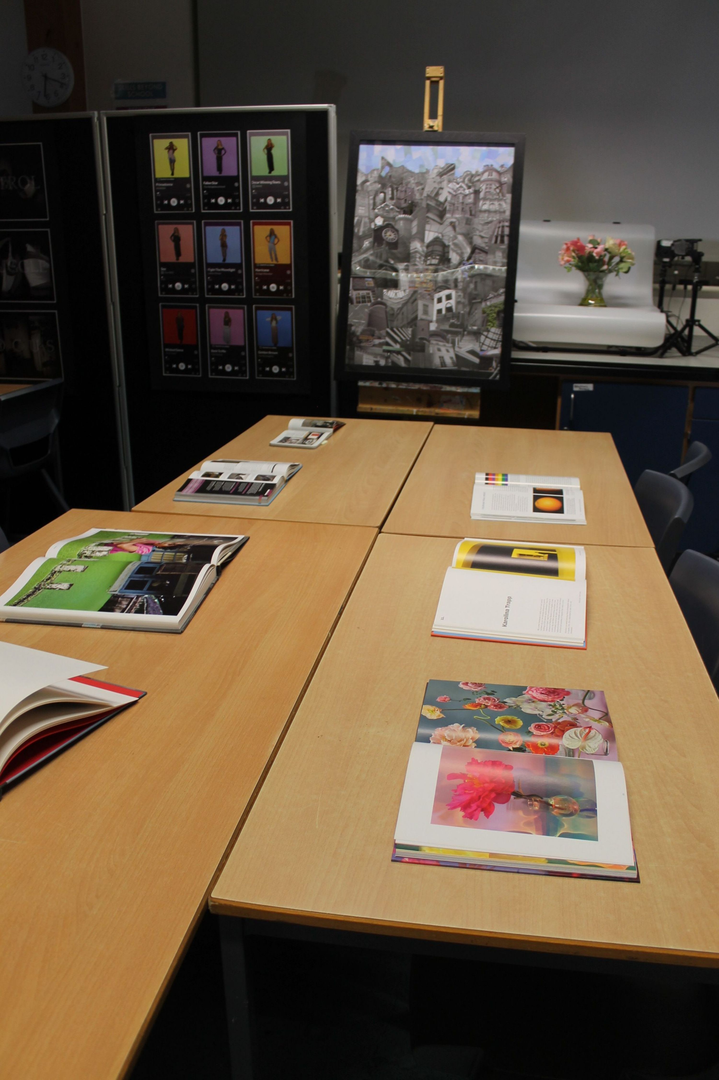 Photograph of an exhibition display presenting student artwork and visual research across wooden panels. Colourful prints, illustrated books, framed studies, a black-and-white monitor, and a grid of labelled graphic designs form a varied gallery-style presentation.
