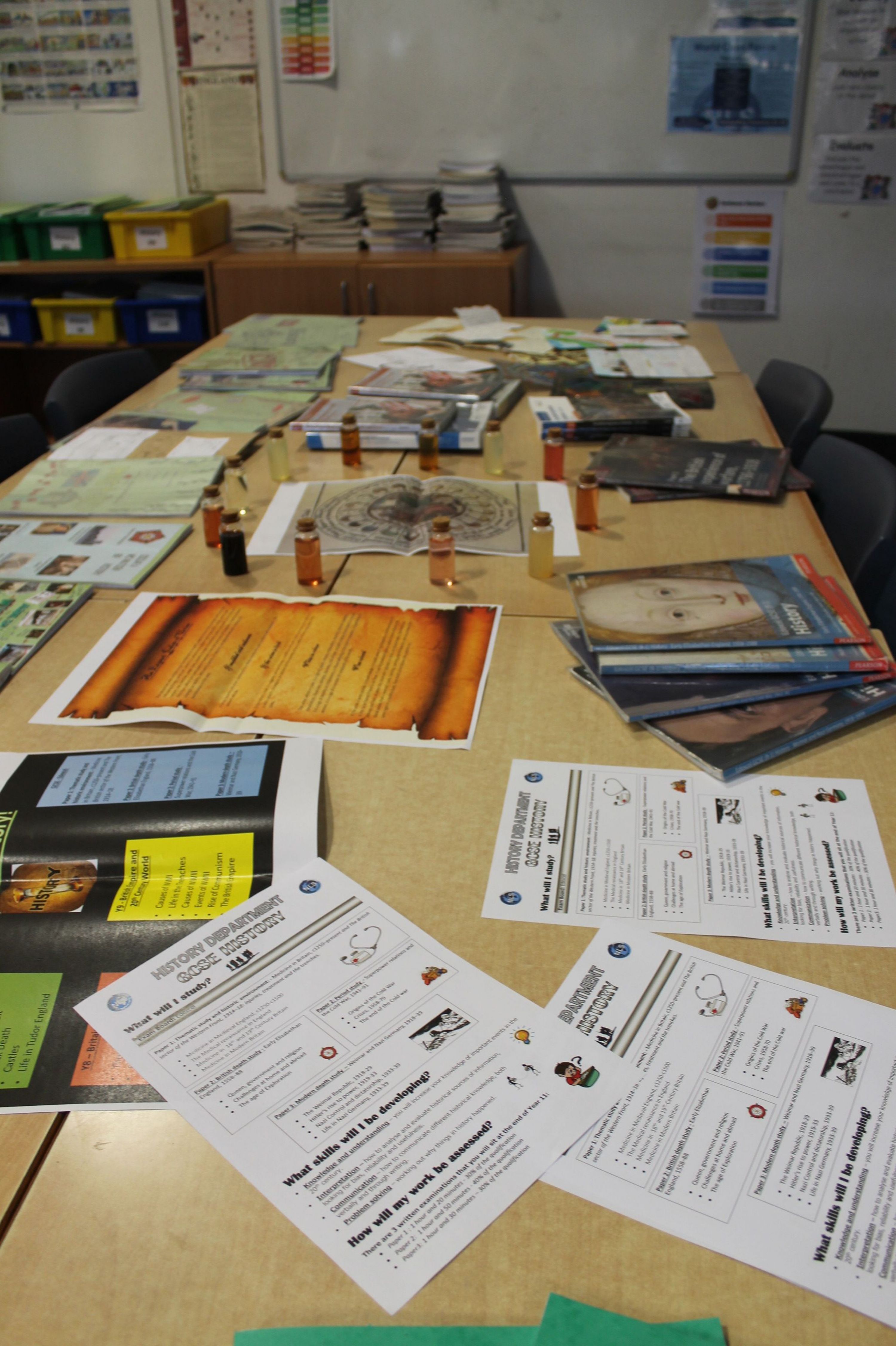 Photograph of a display table covered with books, leaflets, posters and printed guidance materials. Corkboard panels, classroom chairs and wall charts form background, while multilingual-looking handouts, diagrams