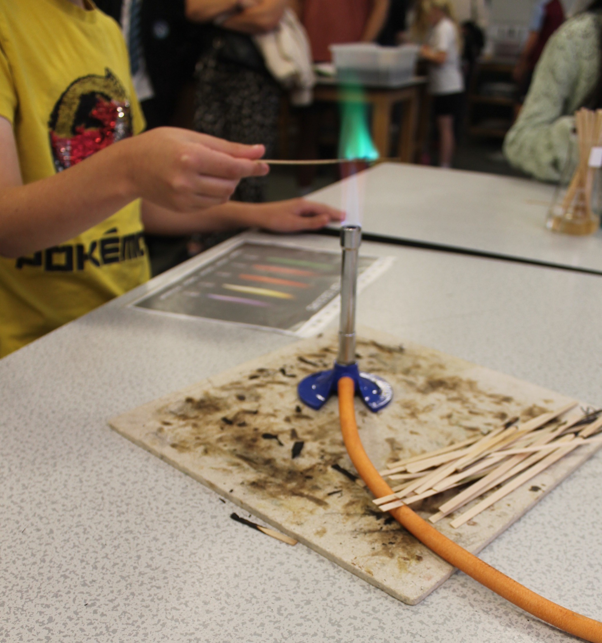 Photograph showing a science experiment at a classroom table, with students operating a metal apparatus, orange hose and wooden sticks on a scorched board. Charred residue, reference sheet and surrounding laboratory materials suggest a heat or combustion investigation.