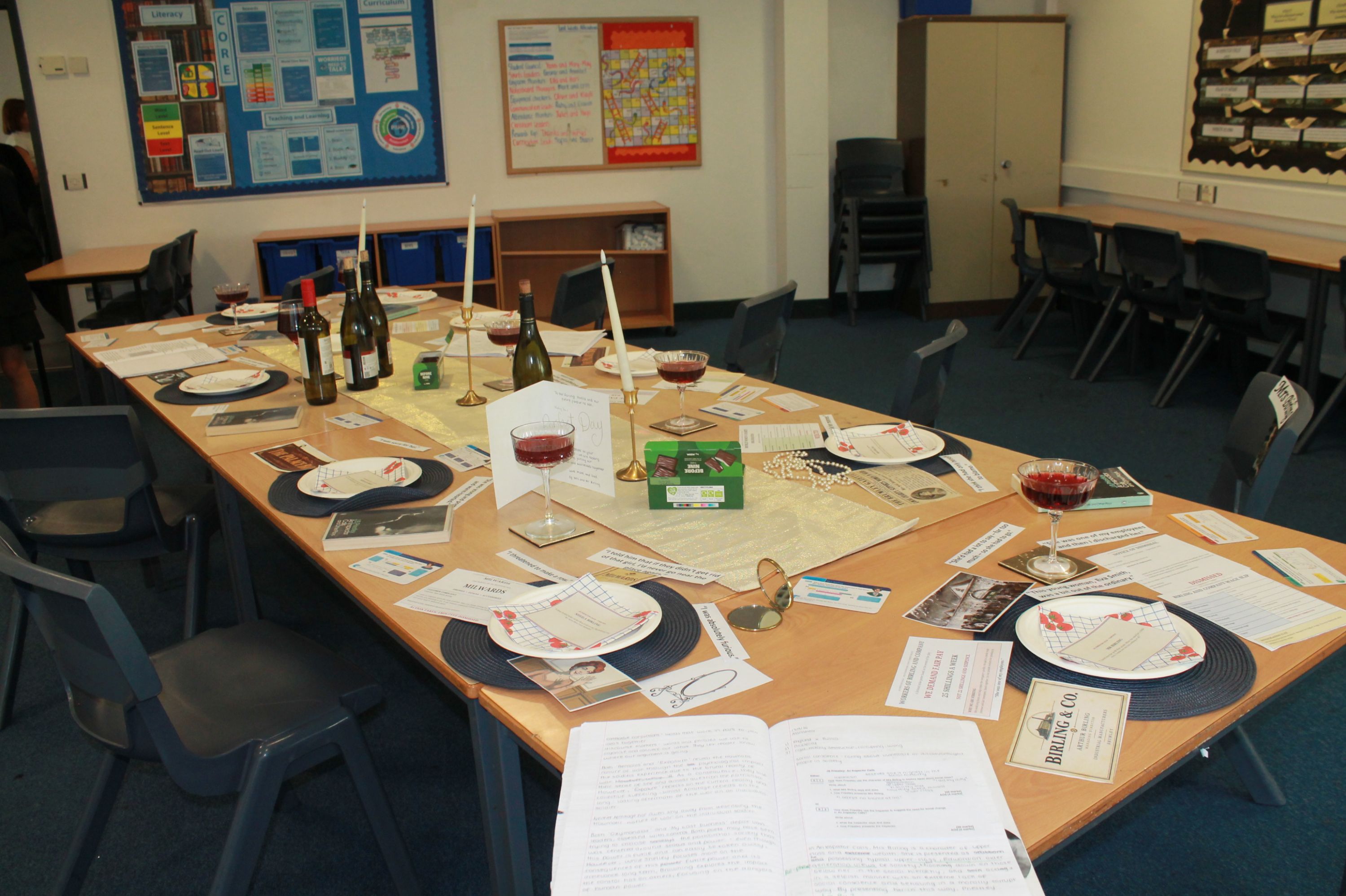 Photograph of a classroom  table arranged for a wine-tasting or food-education session, with bottles, red wine glasses, plates, cutlery, placemats and printed handouts. U-shaped tables hold labelled materials and sample dishes, while educational posters, charts, stacked chairs and storage cupboards fill background.
